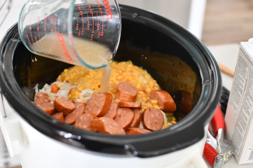 adding broth to slow cooker with sausage and corn.