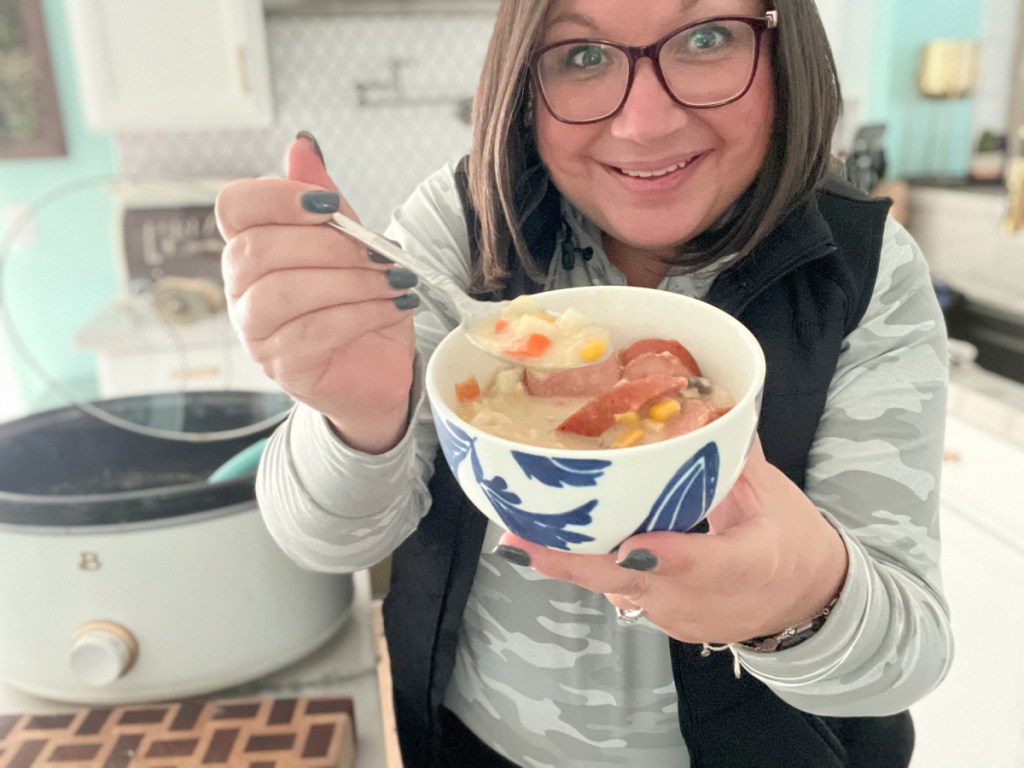 A woman is holding a warm bowl of sausage corn chowder.