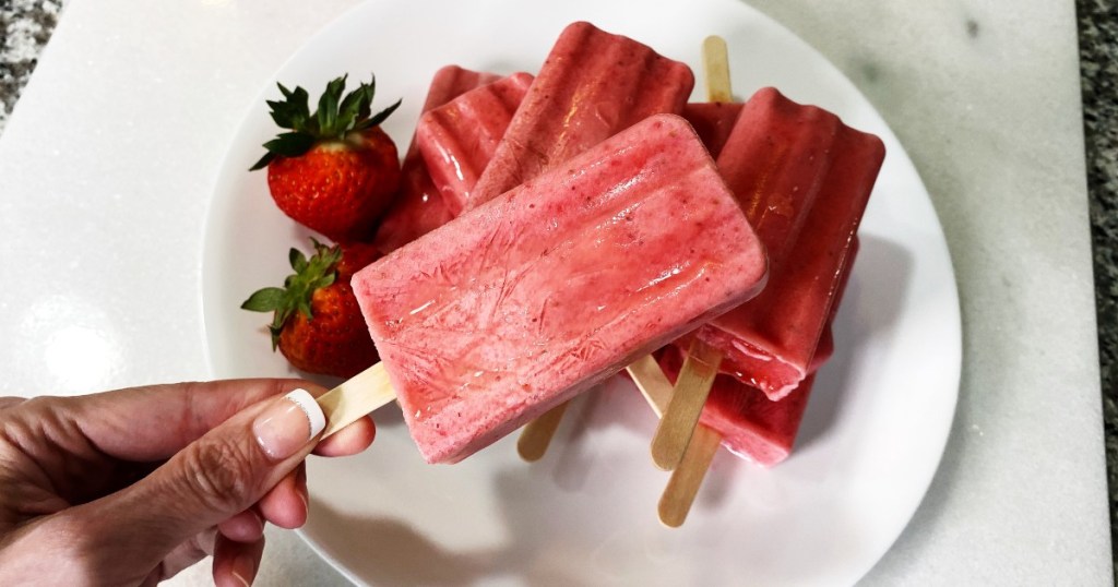 4th of july party ideas - hand holding red strawberry popsicle in front of plate full of popsicles and strawberries