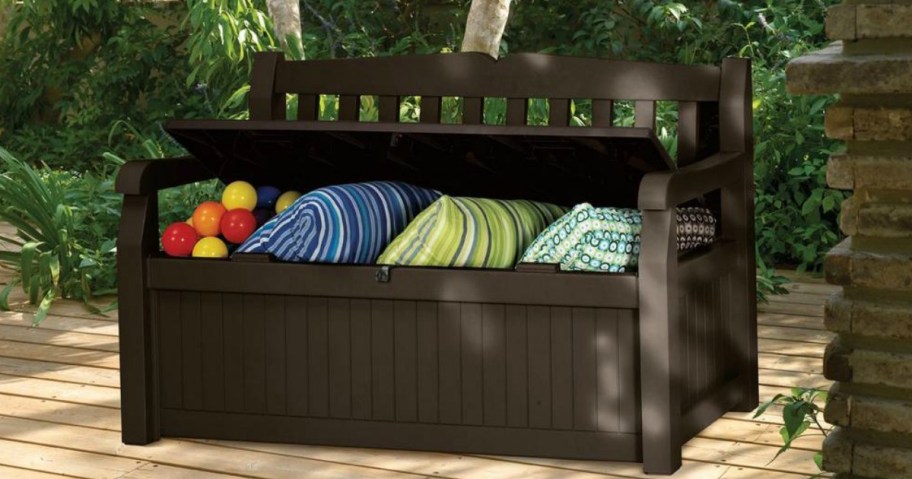 A Keter Solana 70 Gallon Storage Bench Deck Box on a patio filled with pillows and pool toys 