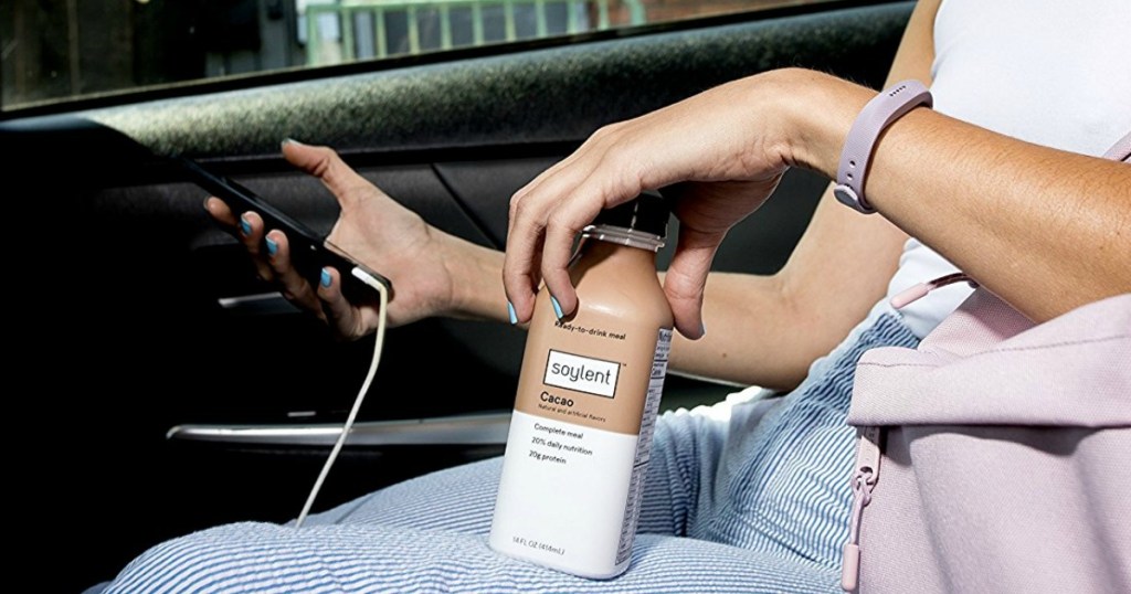 woman holding a Soylent Cacao Bottle while sitting in a car and holding your phone