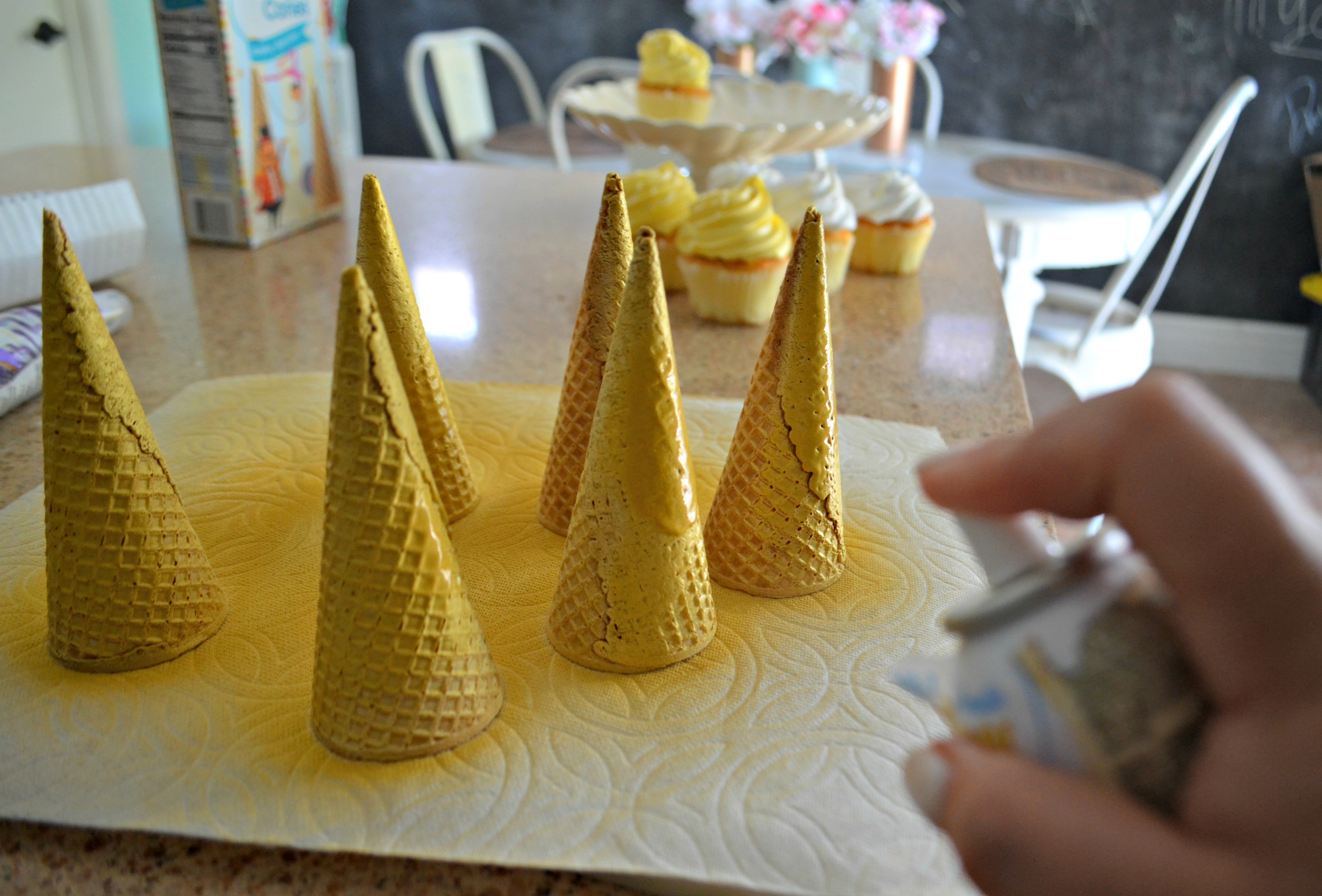 ice cream cones with metallic edible spray on counter with cupcakes in background