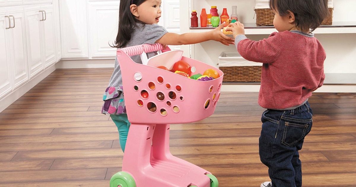 little girl pushing pink little tikes kids shopping cart in kitchen with little boy handing her an item to put in the cart