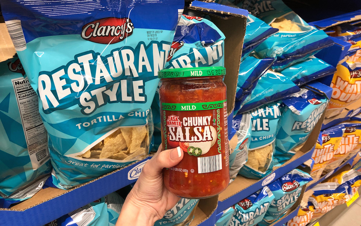 hand holding a jar of salsa next to bags of chips