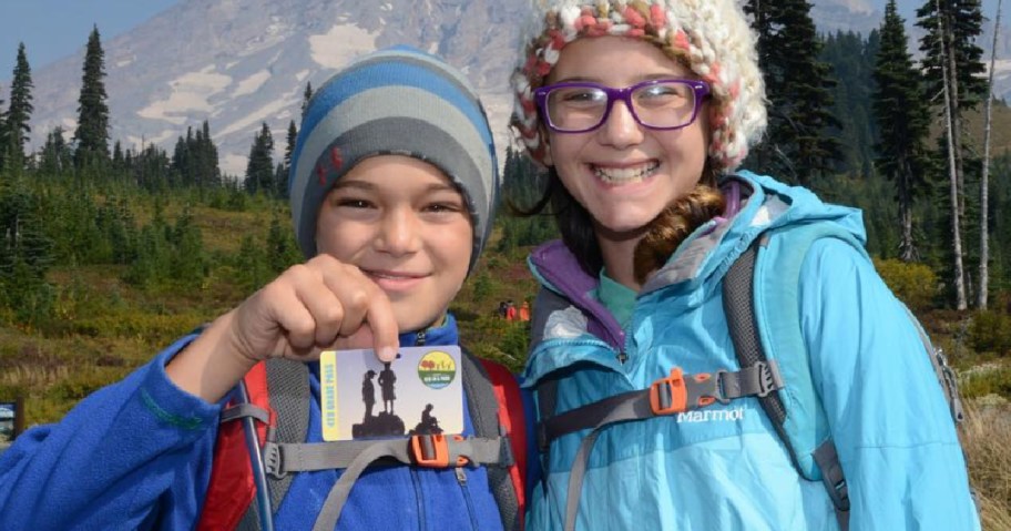 Two kids standing side by side. One is holding up a national park pass.