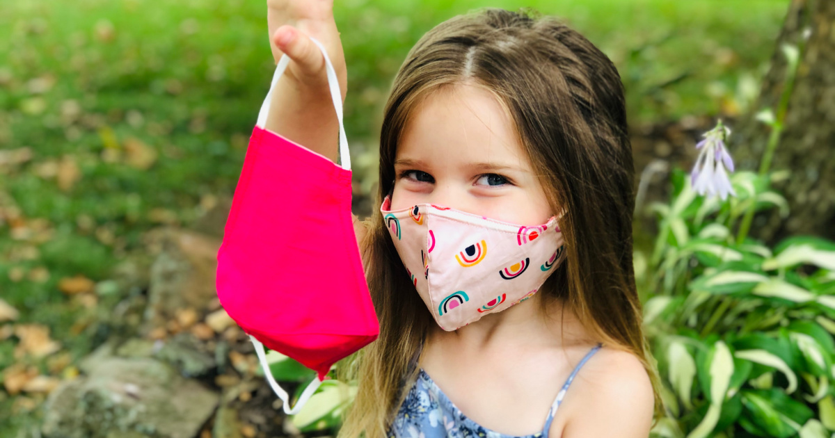 young girl holding hot pink face mask and wearing light pink with rainbows outside