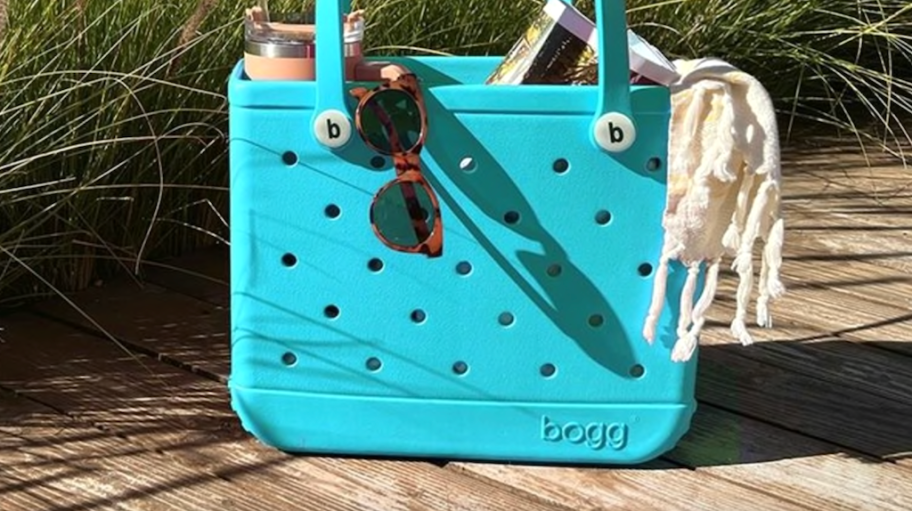 teal blue Bogg Bag sitting on boardwalk at the beach filled with beach items