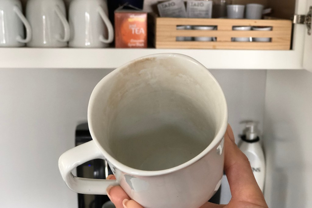 hand holding stained dirty white coffee mug with coffee and mugs in cabinet