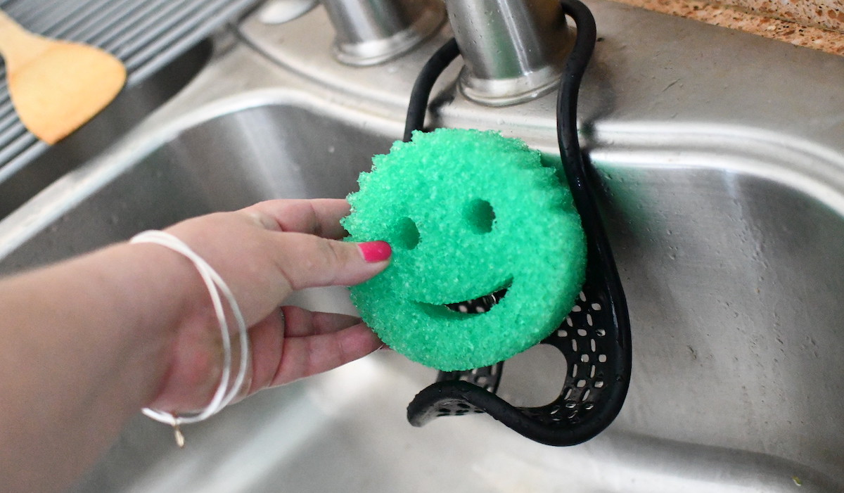 hand holding a green smiley face sponge in stainless steel sink 