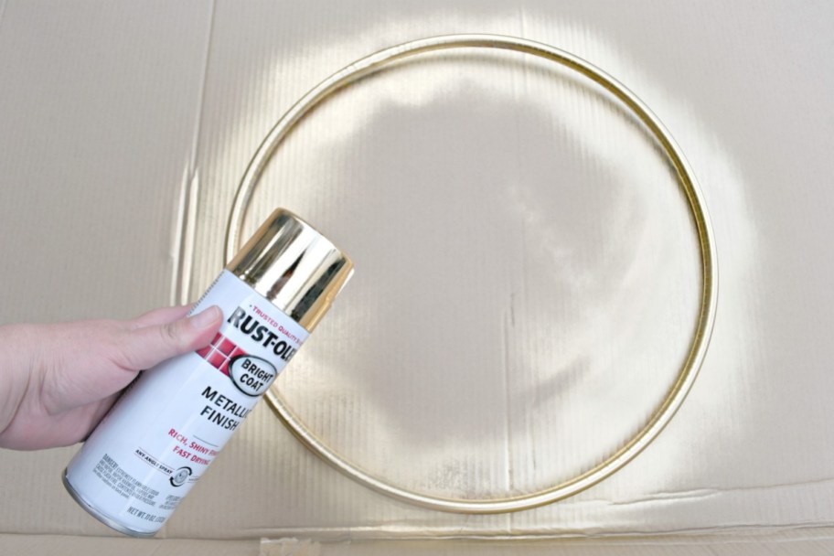 hand holding a can of gold spray paint over painted hula hoop on cardboard