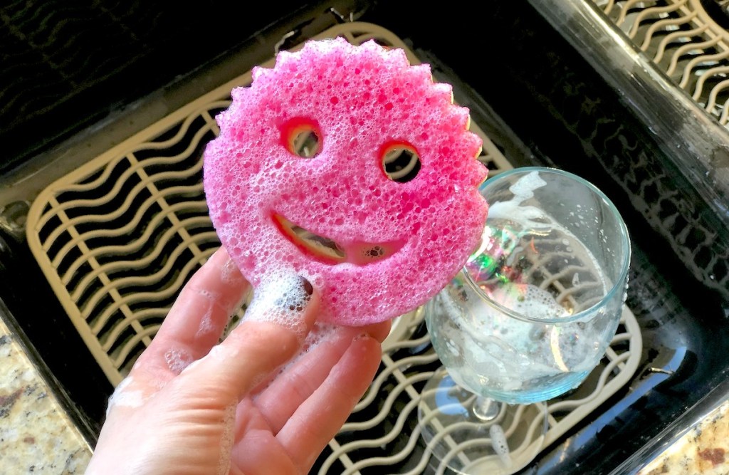 hand holding a pink smiley face sponge with soap suds and wine glass in background