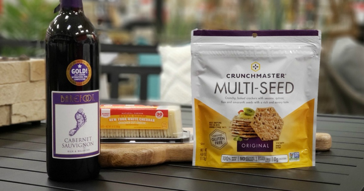 a bottle of wine, a package of sliced cheese sitting on a cutting board, and a bag of Crunchmaster crackers