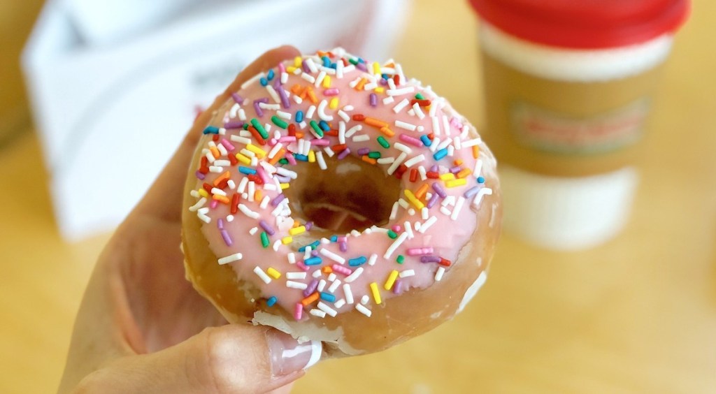 hand holding a pink glazed donut with rainbow sprinkles and coffee in background