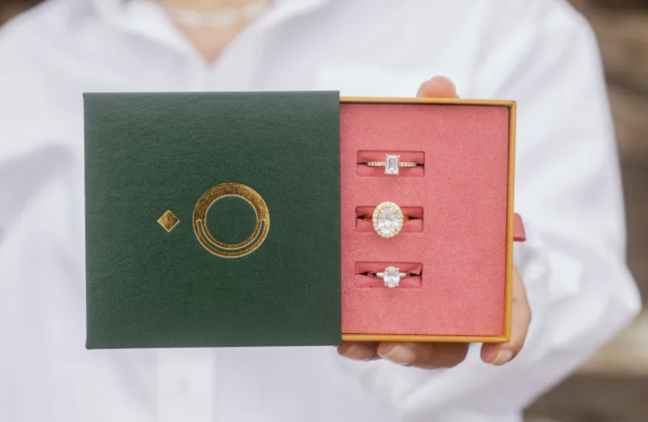hand holding a green and pink box with three diamond rings inside