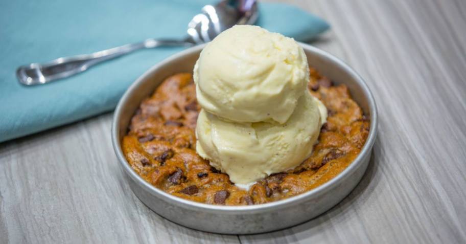 deep dish chocolate chip cookie with two scoops of vanilla ice cream on top with spoon and blue napkin