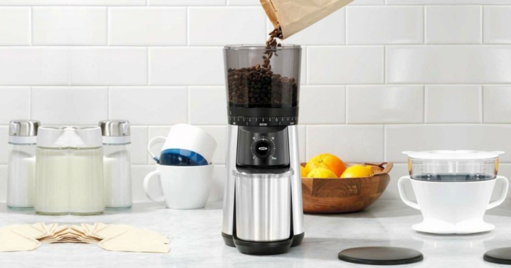 Coffee beans being poured into OXO BREW Conical Burr Coffee Grinder on countertop in kitchen