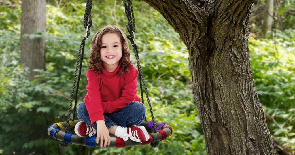 little girl sitting on sorbus swing which is hanging from a tree with trees in background