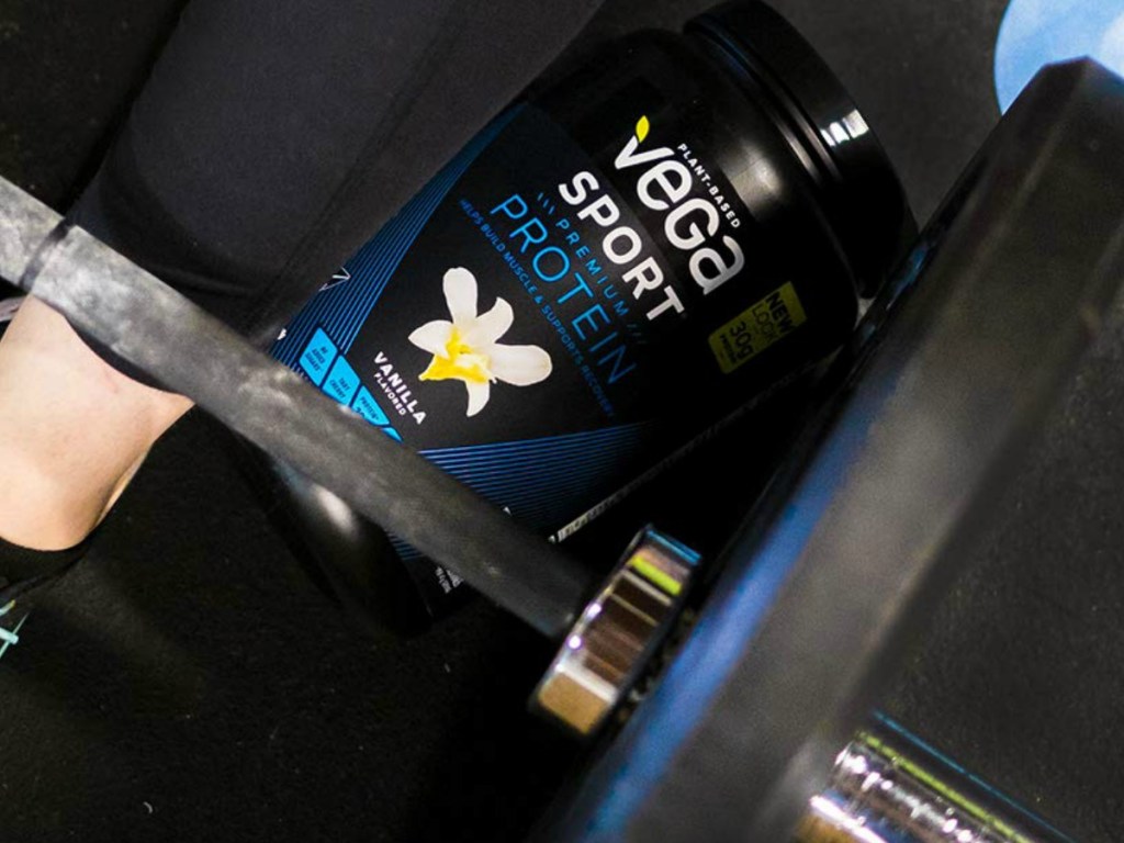 vega sport protein powder in black canister under weight bench