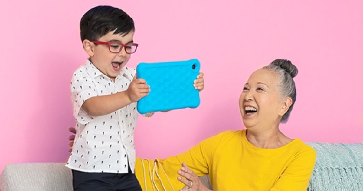 boy holding blue amazon kids fire table while standing on a couch with older woman