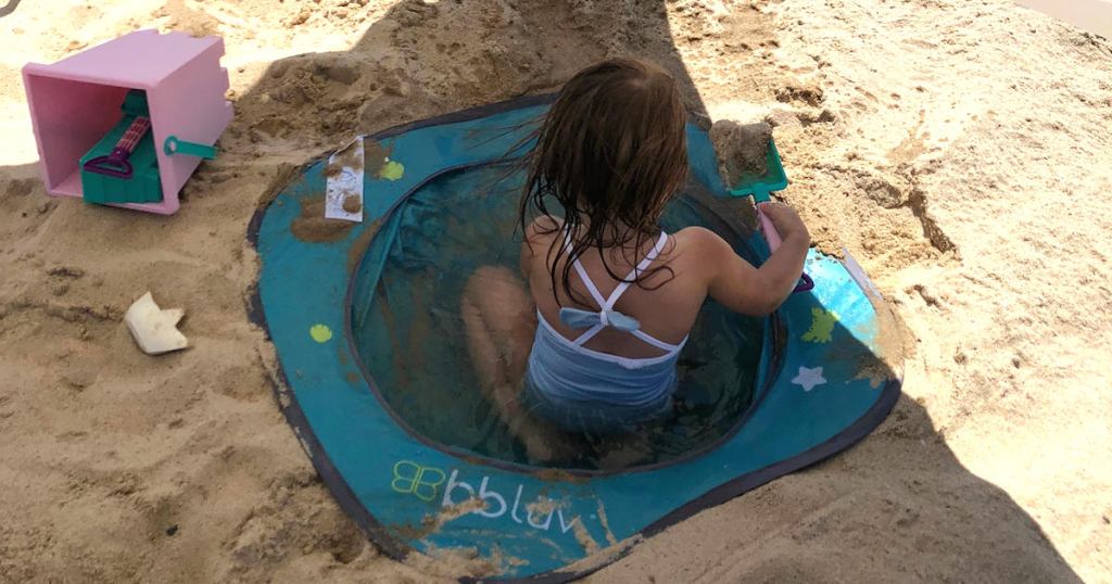 baby girl wearing a blue bathing suit sitting in blue pool in sand