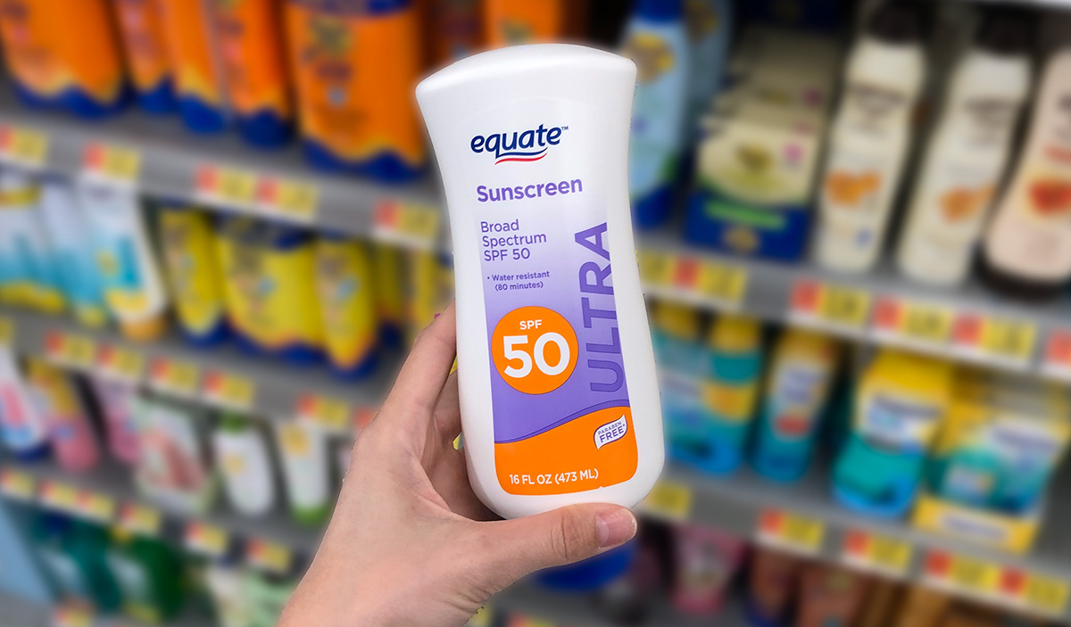 Hand holding a bottle of equate sunscreen with various brands and bottles on store shelf in background