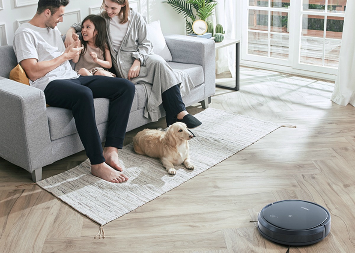 family sitting on couch while ECOVACS DEEBOT 500 Robotic Vacuum cleans wood floor and dog on rug