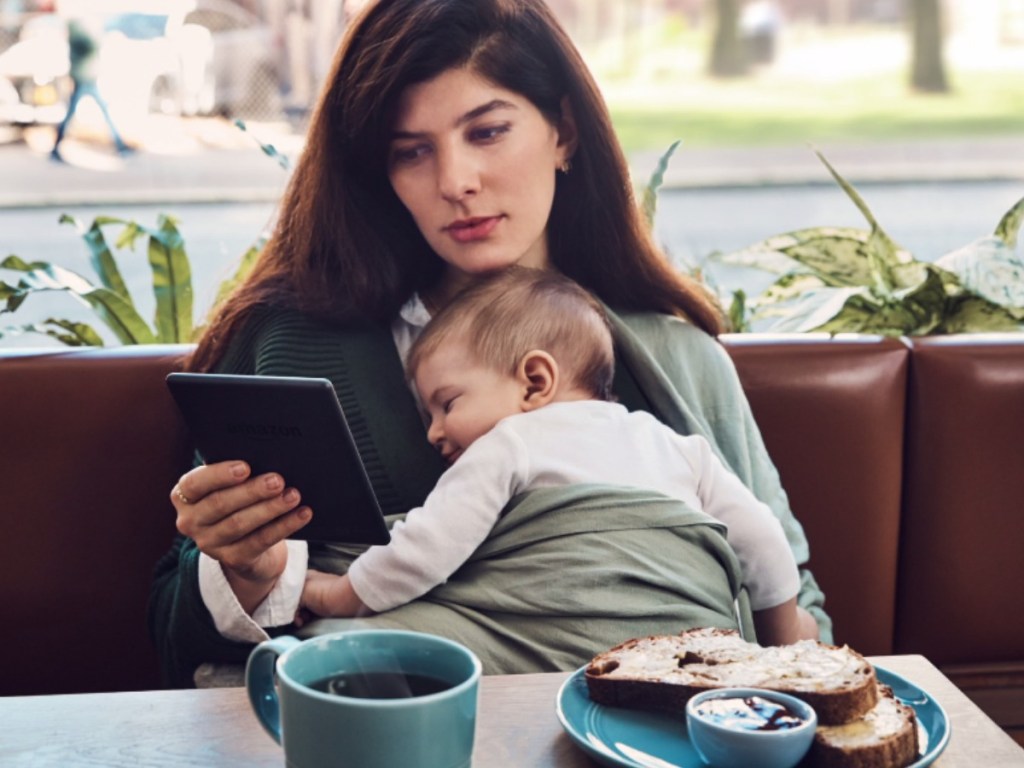woman holding sleeping baby at breakfast table reading ebook