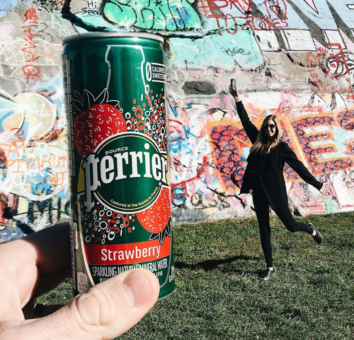 Woman next to can of strawberry Perrier in front of graffiti wall