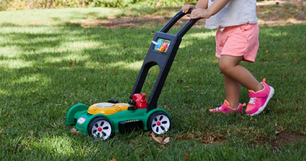 Green toy mower being pushed by girl through grass yard