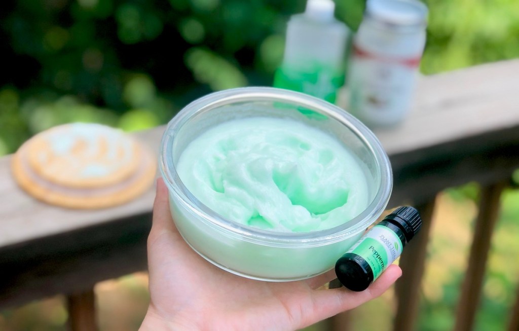 Hand holding a clear glass bowl with green whipped lotion inside and small bottle of peppermint essential oil