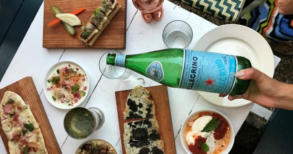 hand pouring San Pellegrino sparkling water into a glass surrounded by food