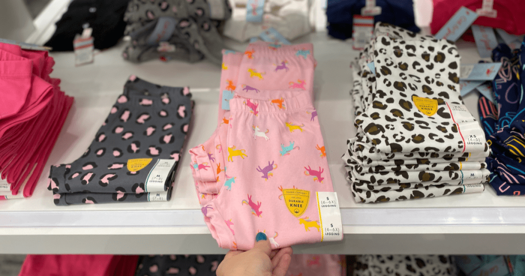 hand holding pink unicorn leggings with other leggings on shelf behind it