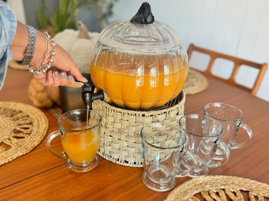 glass pumpkin dispenser filled with apple cider, being dispensed into pretty glasses on a table with fall decor