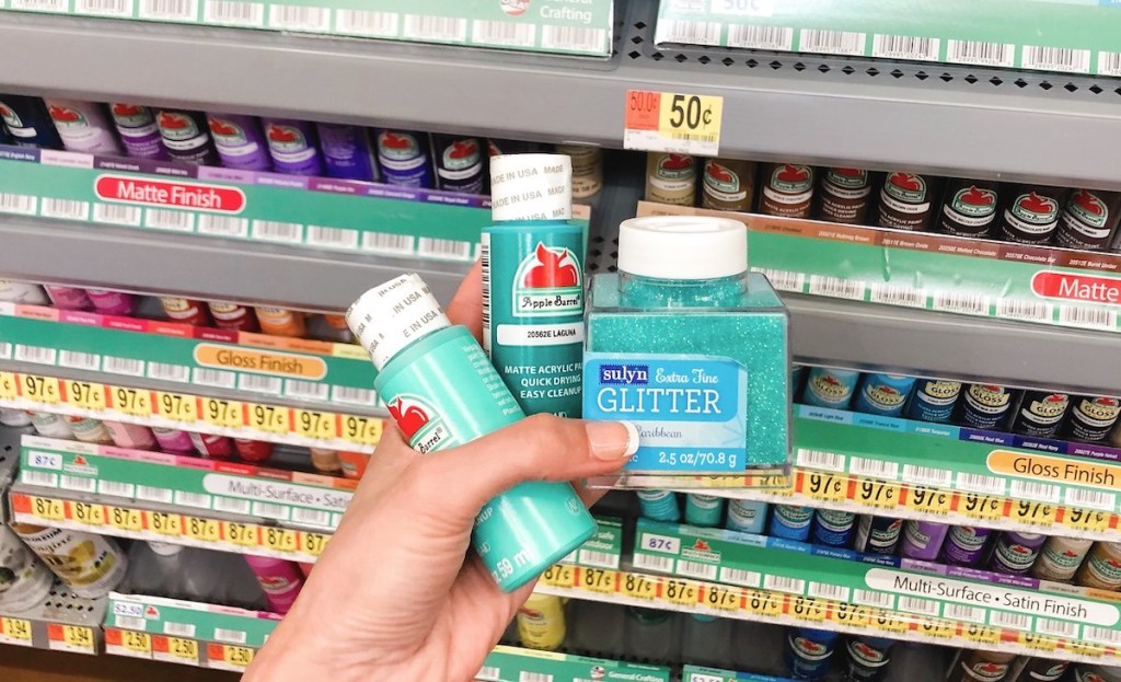 hand holding teal colored paint and glitter in front of store craft aisle