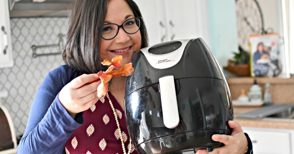 woman holding black air fryer in one hand and bacon in the other