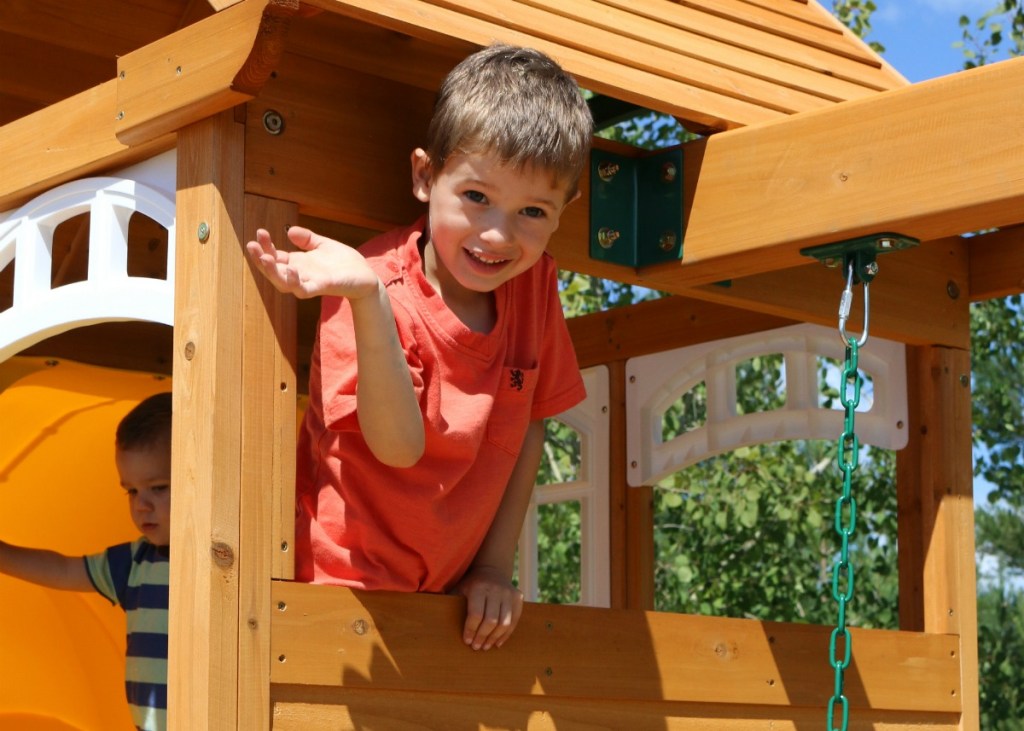 boy waving from the top of a wooden outdoor playset