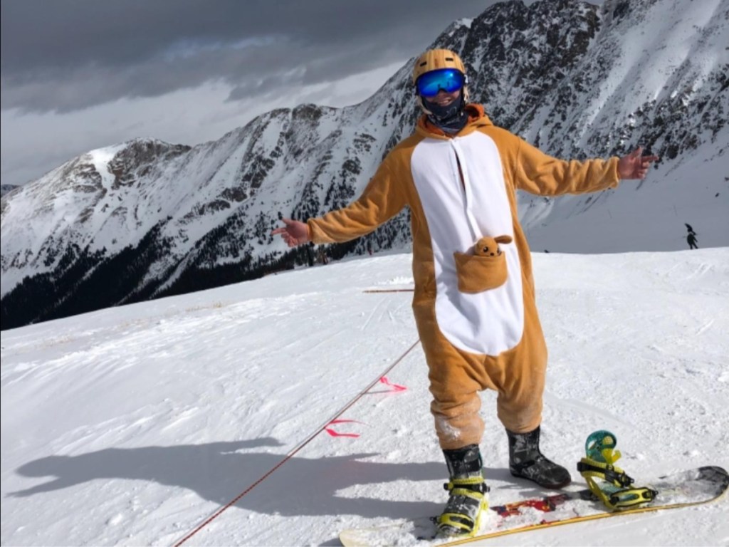 man in kangaroo costume snowboarding with mountains behind him