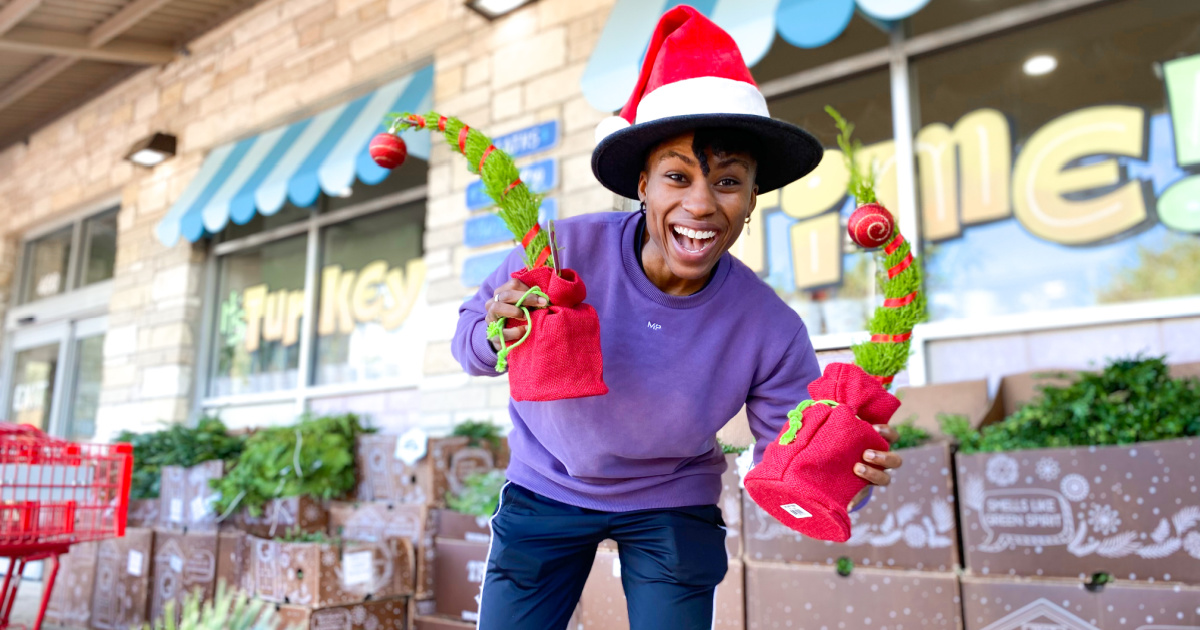 smiling woman wearing a Santa hat and holding two Grinch Christmas Trees outside of a Trader Joe's store