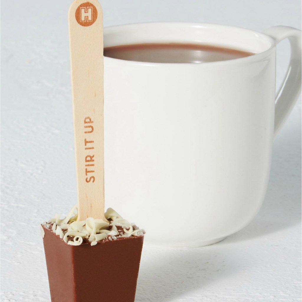 Popsicle stick with solid chocolate at the end next to a white mug full of cocoa
