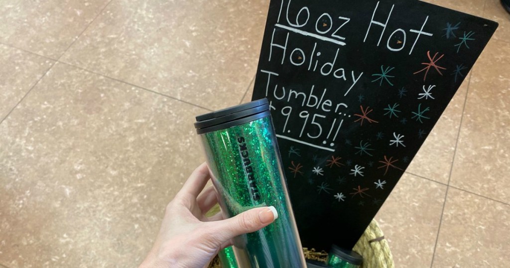 woman holding a green Starbucks holiday tumbler in front of a sale sign