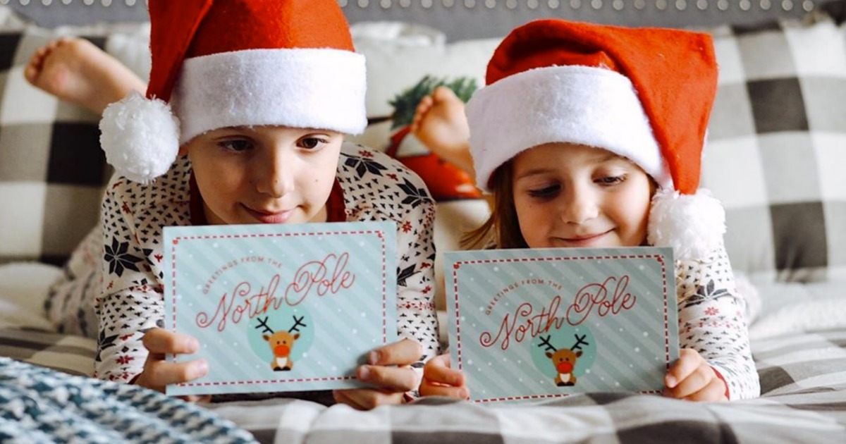 Two kids holding postcards from The North Pole and wearing Santa hats