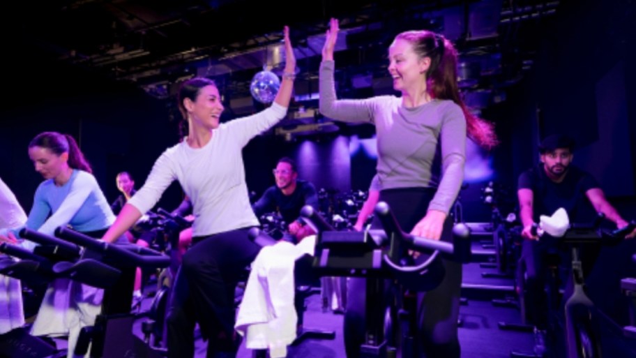 2 women high 5 ing on exercise cycles in a class