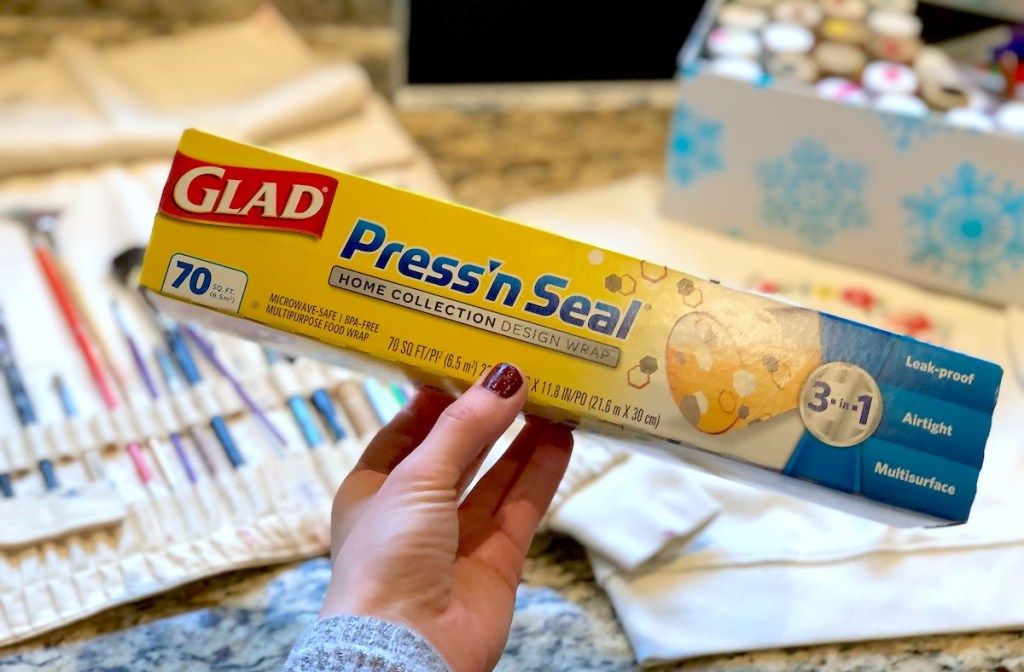 hand holding a package of press and seal plastic wrap with paint supplies in the background