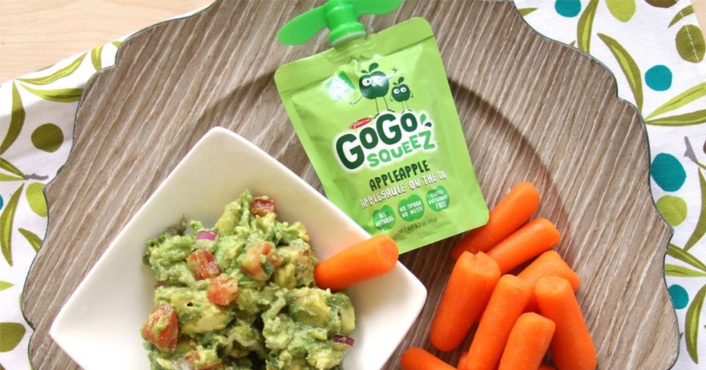 Applesauce pouch, guacamole, and carrots on a table.