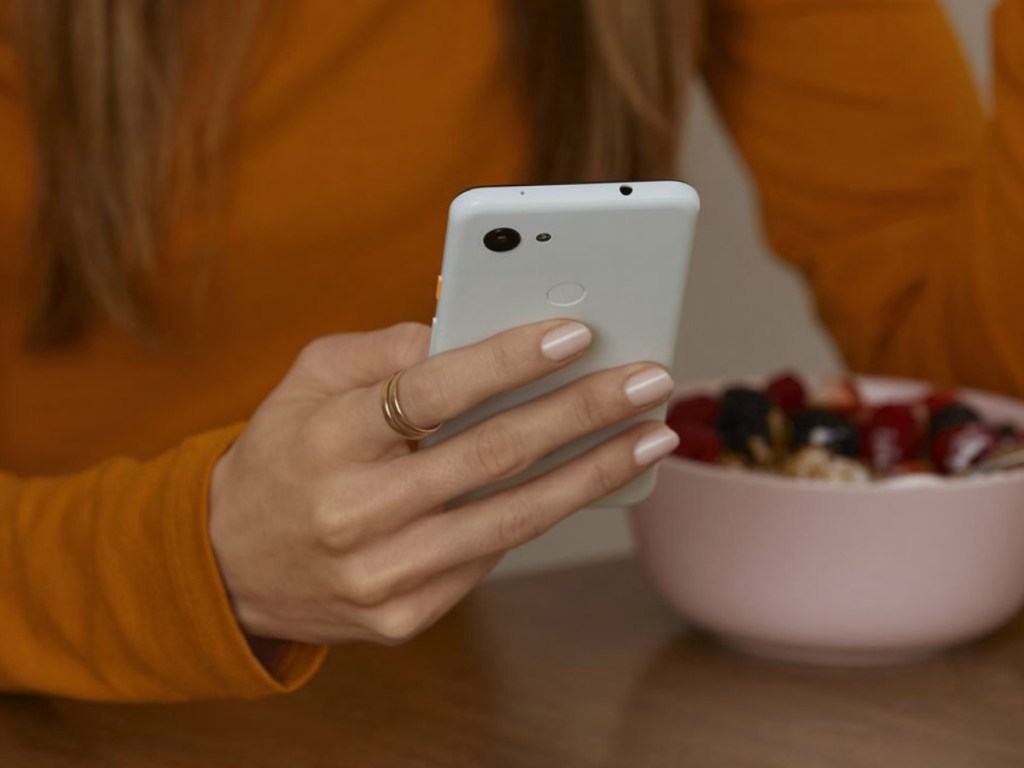 Woman holding a white smartphone with a table and a bowl of fruit