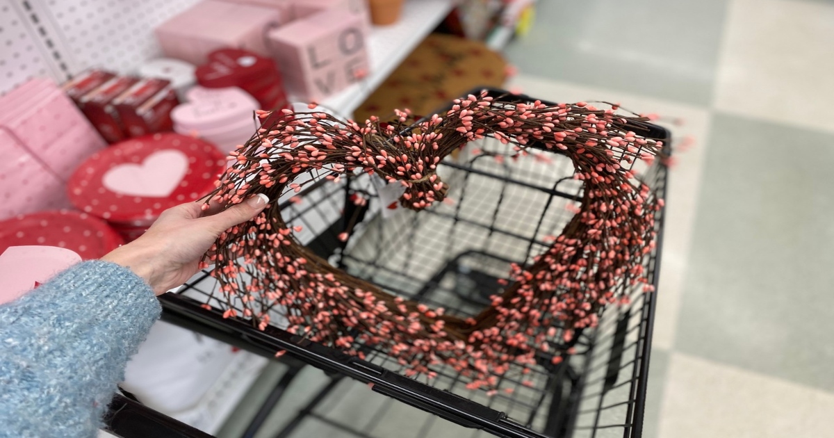 Heart wreath, held by a woman's hand over top of a cart, in store with other valentines items on a rack behind the cart