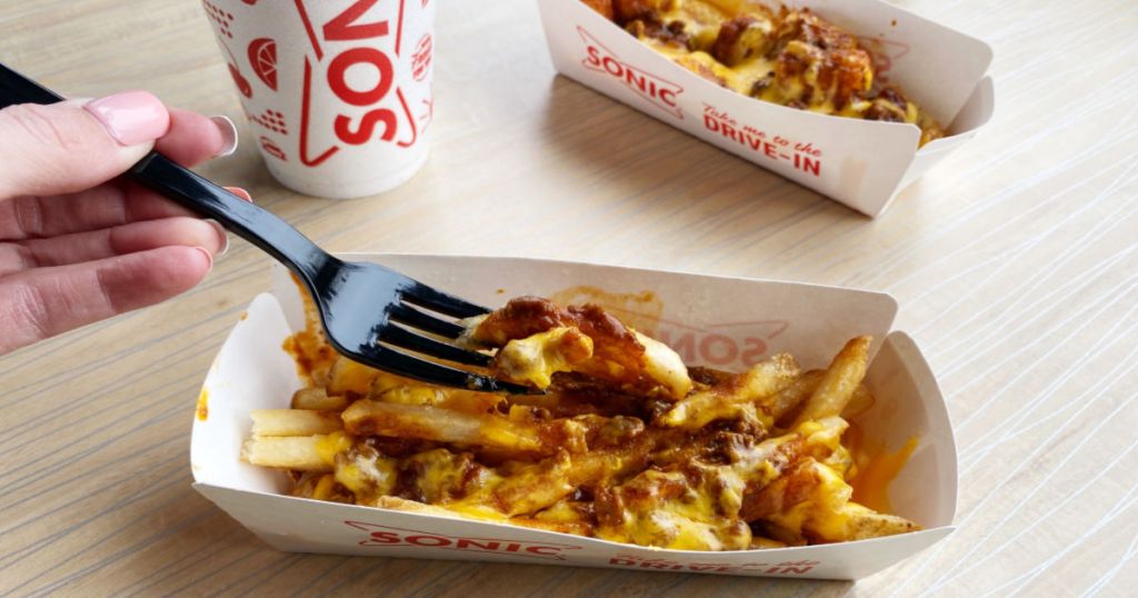 woman holding fork with sonic chili cheese fries on it with chili cheese tots and drink cup in the background