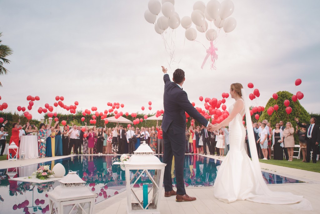 married bride and groom standing in front of wedding party with pool and tons of red and white balloons wedding ideas on a budget