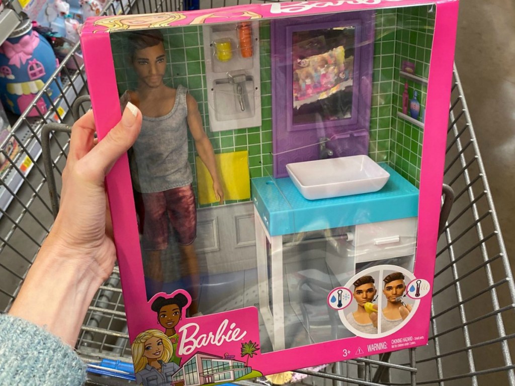 hand holding box with Barbie boy and bathroom set in front of grocery cart