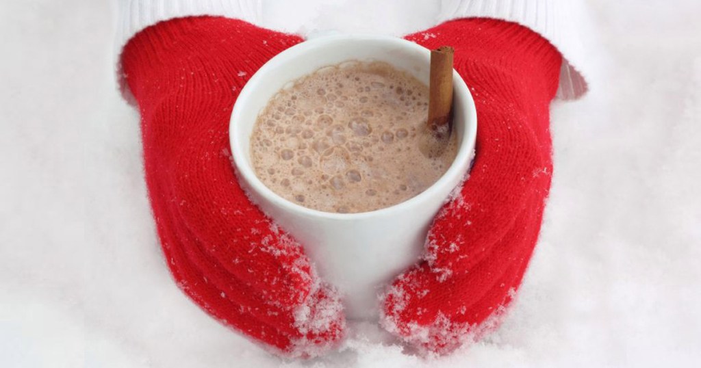 Hands wearing red mittens holding a cup of hot cocoa in the snow